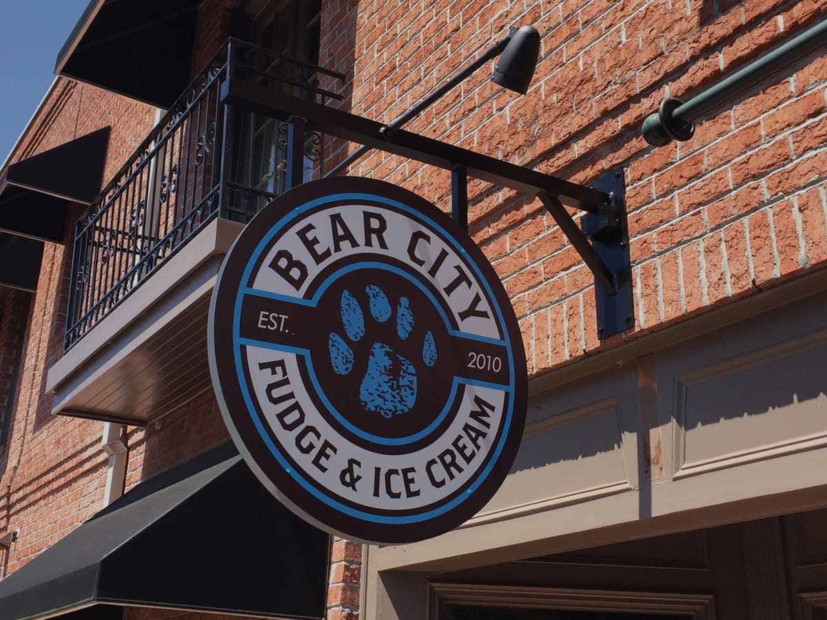 Bear City Fudge & Ice Cream round hanging blade sign on an iron bracket against brick