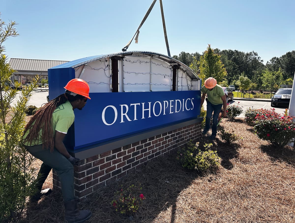 Crew in hard hats setting an orthopedics sign cabinet onto its brick monument base with a crane