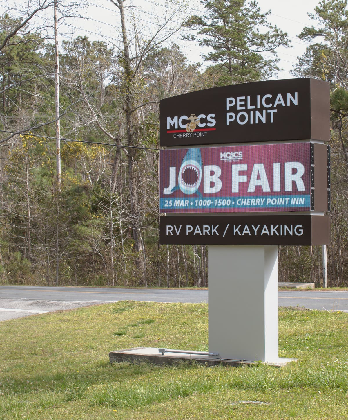 MCCS Pelican Point pylon with LED message board beside a wooded base road at dusk