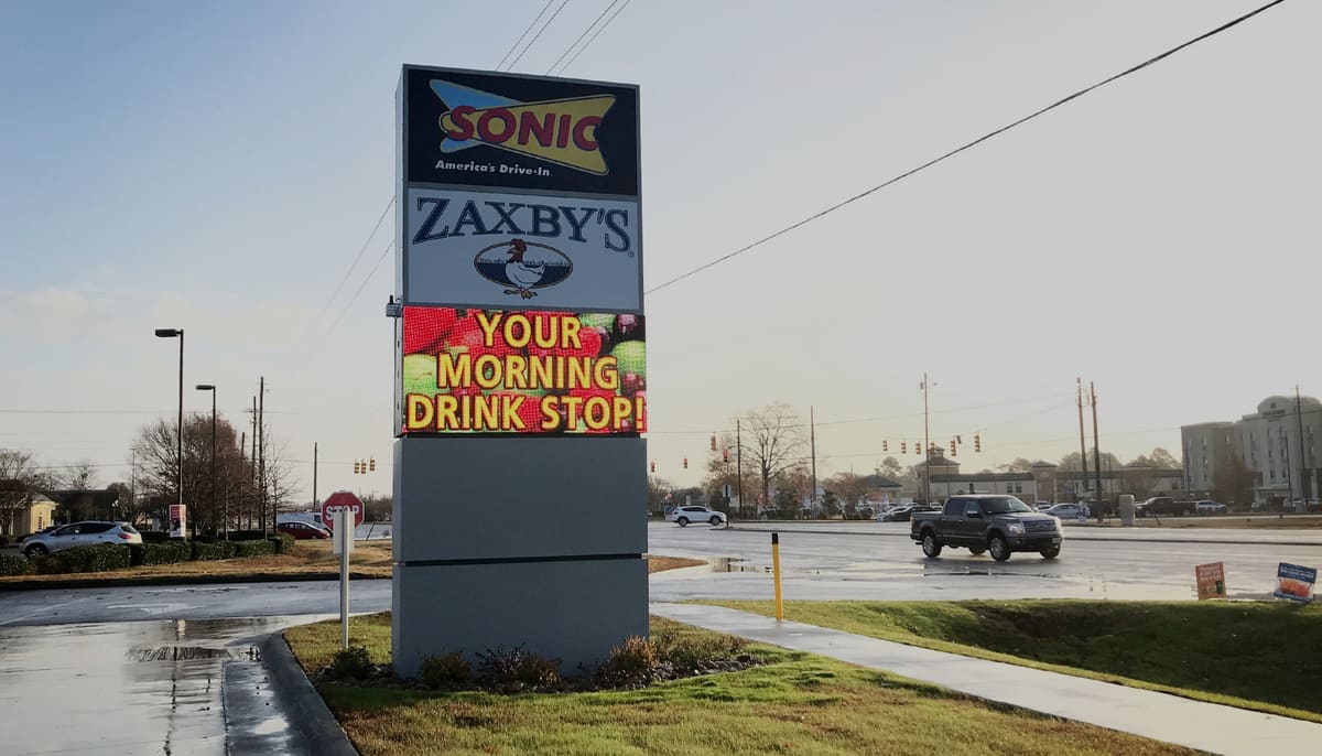 Shared Sonic and Zaxby’s pylon with LED message board over a wet parking lot in morning light
